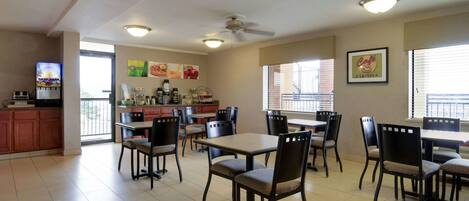 A spacious dining area with multiple tables and chairs, a kitchen counter with coffee machines, and a television displaying a program.
