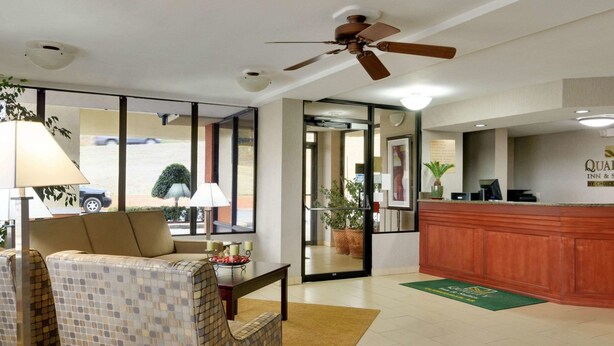 Un hall de hotel con una mesa de recepción, un sofá, una silla, un ventilador de techo y un ventanal con vistas al exterior.