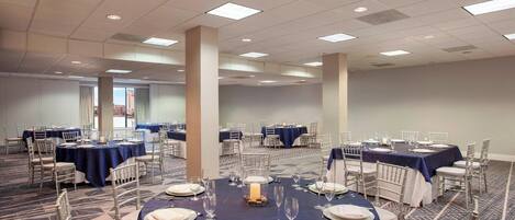 A large conference room with multiple round tables, each set with white tablecloths, plates, and glassware.