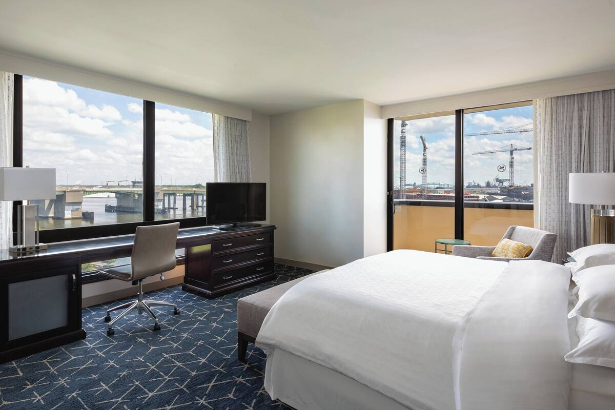 A hotel room with a large bed, a desk with a chair, a television, and a view of a construction site through large windows.