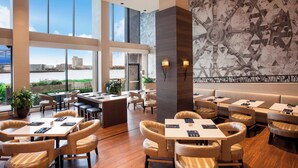 A modern hotel lobby with high ceilings, large windows, and a view of the cityscape. There are multiple seating areas with tables and chairs, and a large abstract mural on the wall. - Sheraton Norfolk Waterside Hotel (Norfolk)