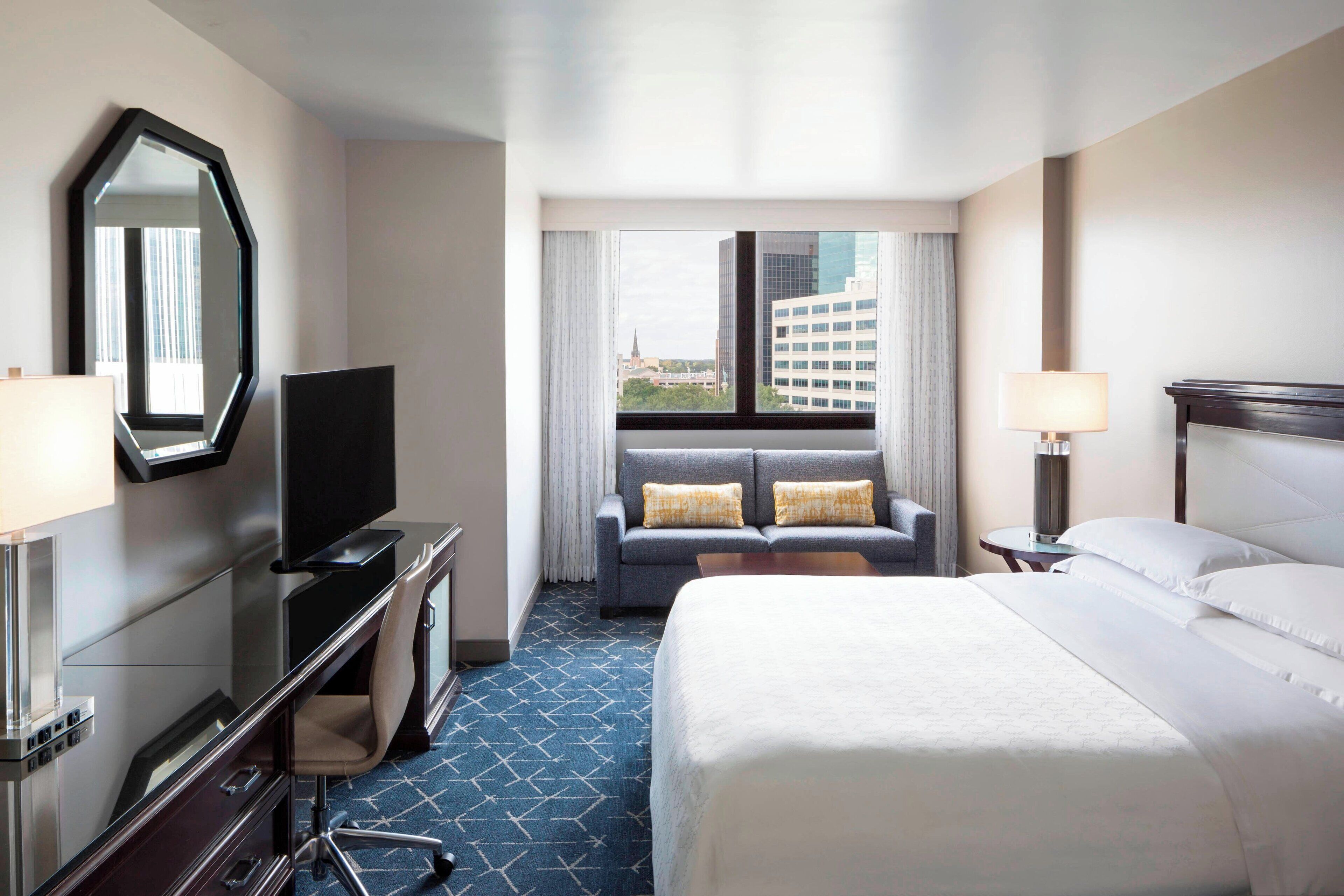 A hotel room with a large bed, a desk with a TV, a sofa, a window with curtains, and a lamp.