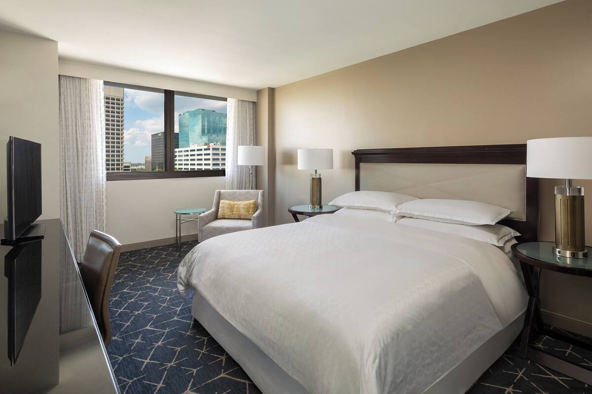 A hotel room with a large bed, two bedside tables with lamps, a chair, a desk with a TV, and a window with a city view.