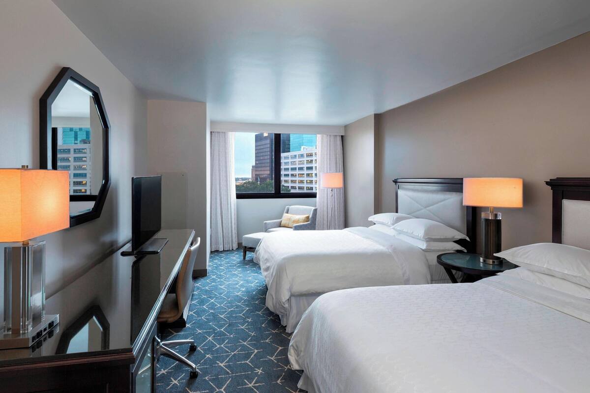A hotel room with two beds, a desk, a chair, a TV, and a view of buildings through the window.