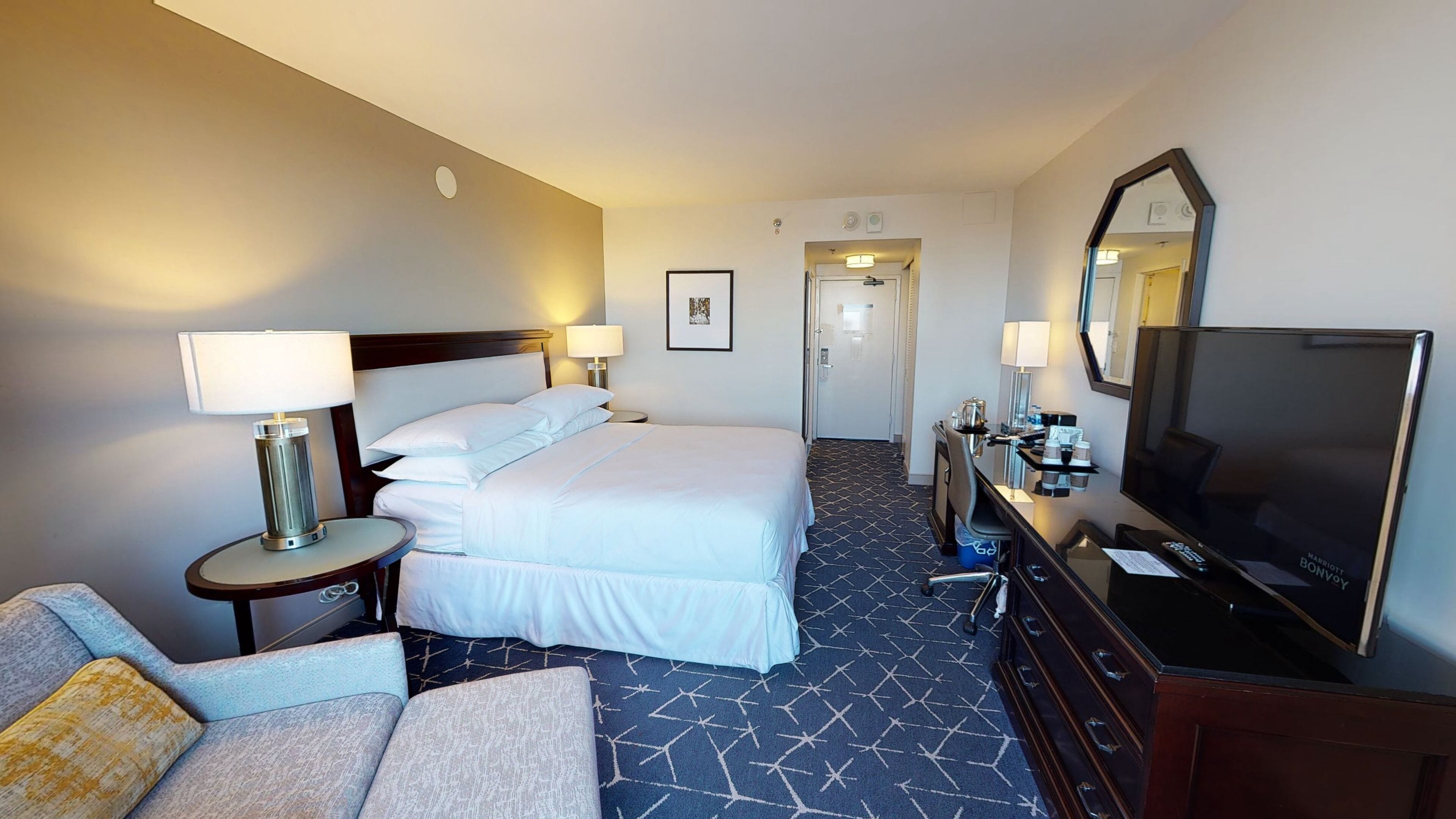 A hotel room with a bed, two bedside tables with lamps, a TV, a dresser, and a sofa.