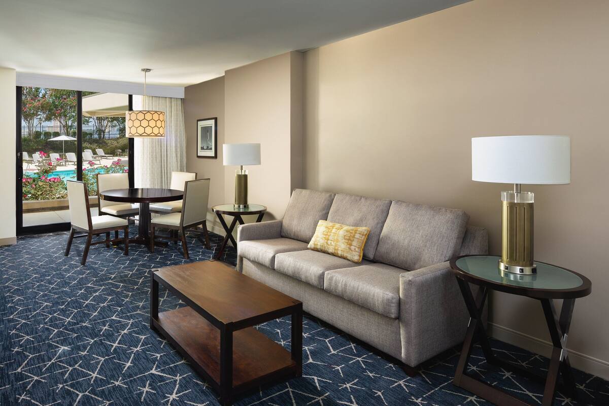 A hotel room with a sofa, a coffee table, a dining table with chairs, a lamp, and a view of a pool and garden.