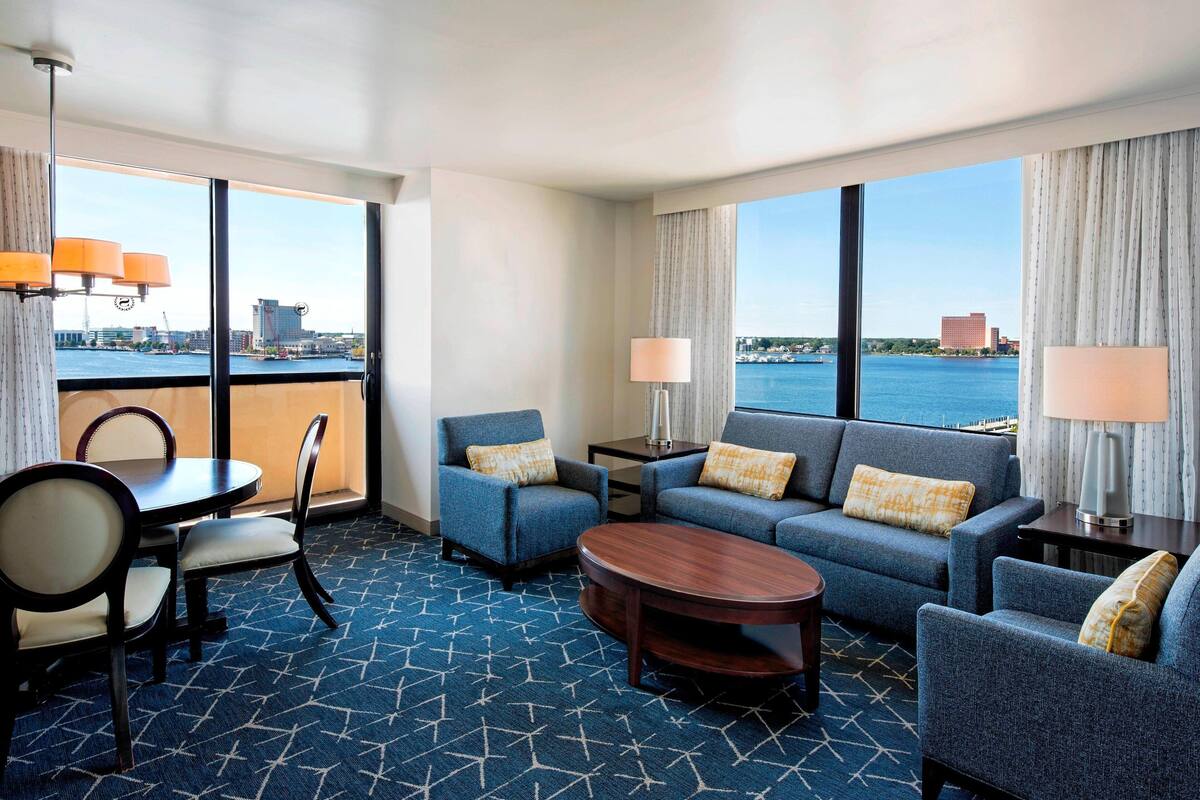 A hotel room with a large window, a sofa, two armchairs, a dining table with chairs, and a small coffee table.