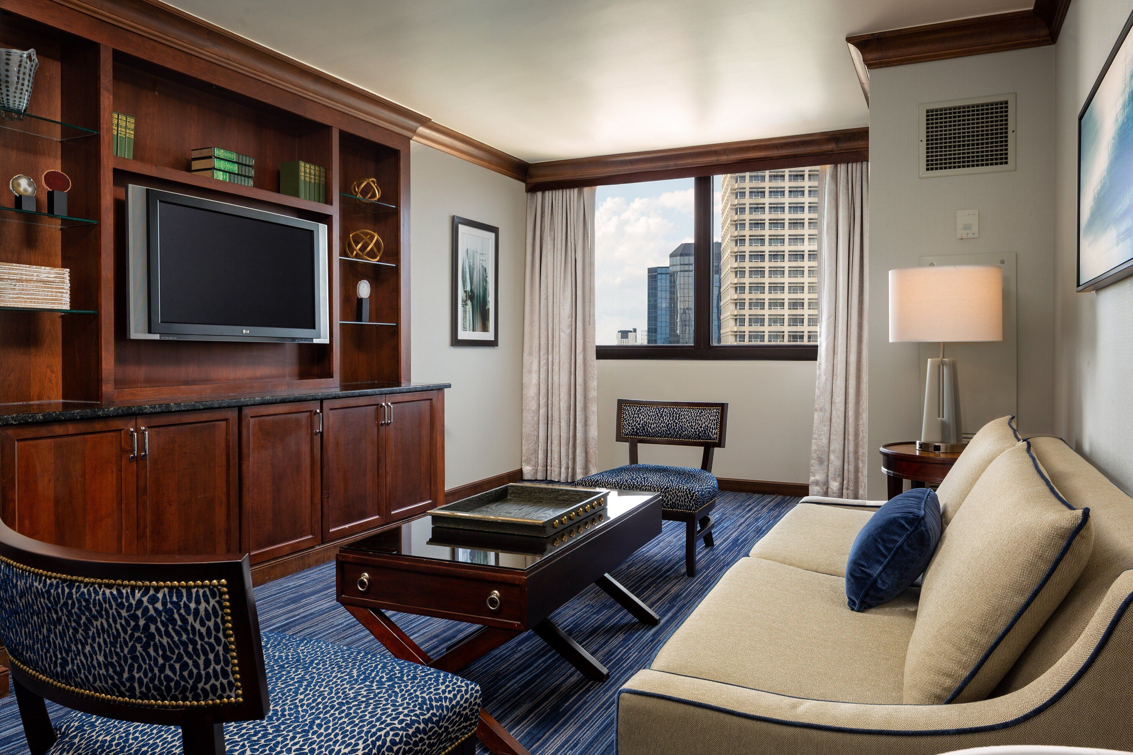 Habitación de hotel con televisión de pantalla plana, armarios de madera, sofá, sillón estampado y vista a la ciudad desde la ventana.