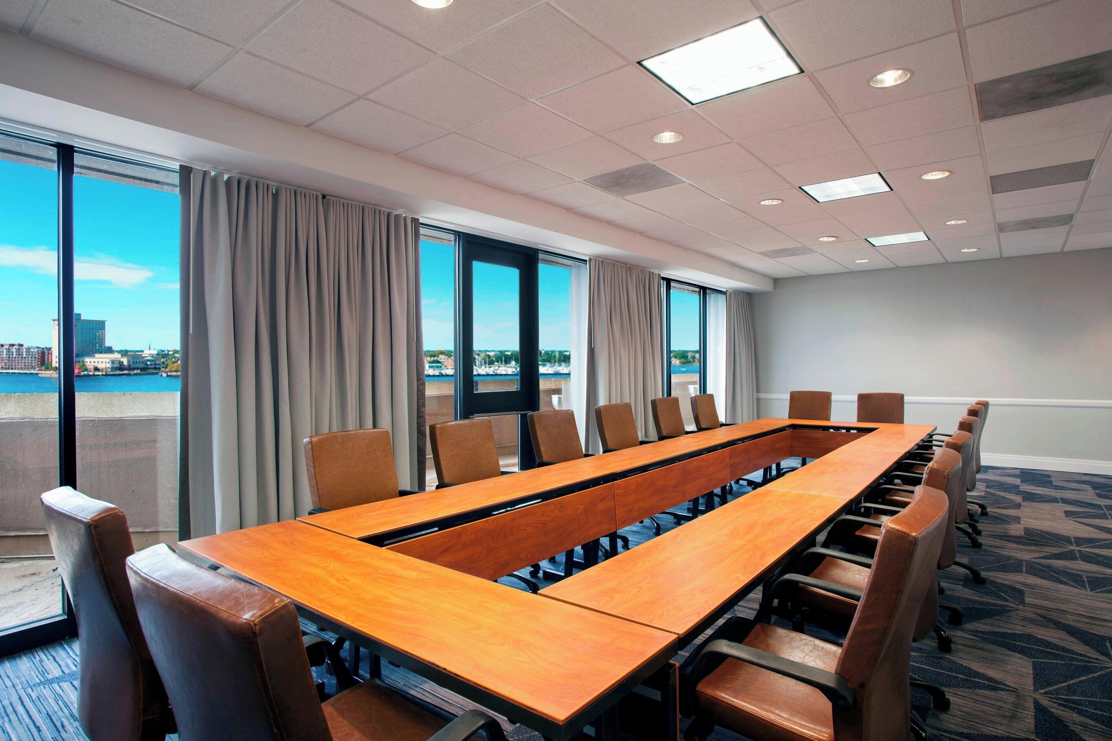Un salón de conferencias con una larga mesa y sillas, ventanas amplias con cortinas y vista a la ciudad.