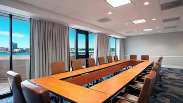 Un salón de conferencias con una larga mesa y sillas, ventanas amplias con cortinas y vista a la ciudad.