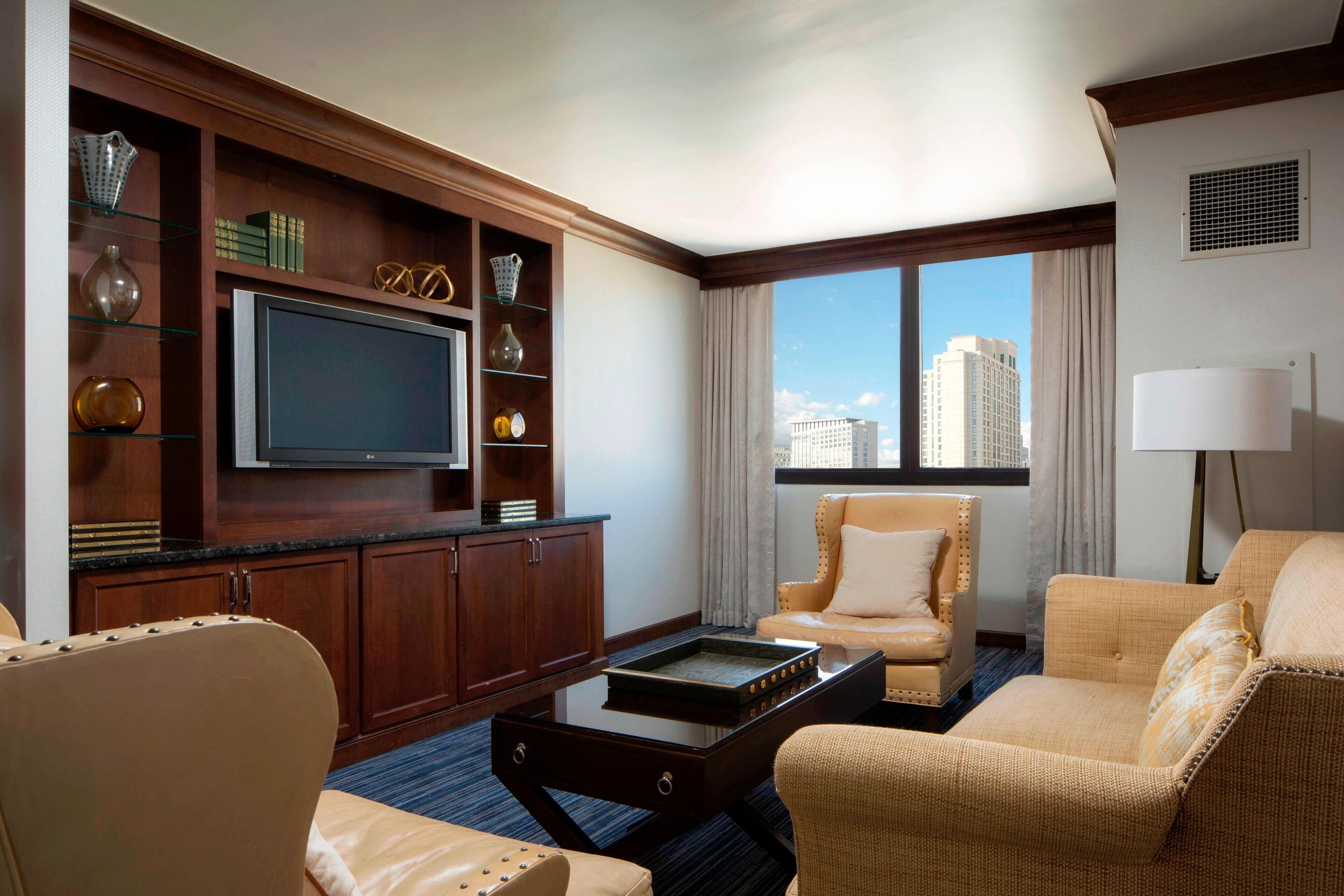 A hotel room with a flat-screen TV, a bookshelf, a sofa, a chair, and a large window with a city view.