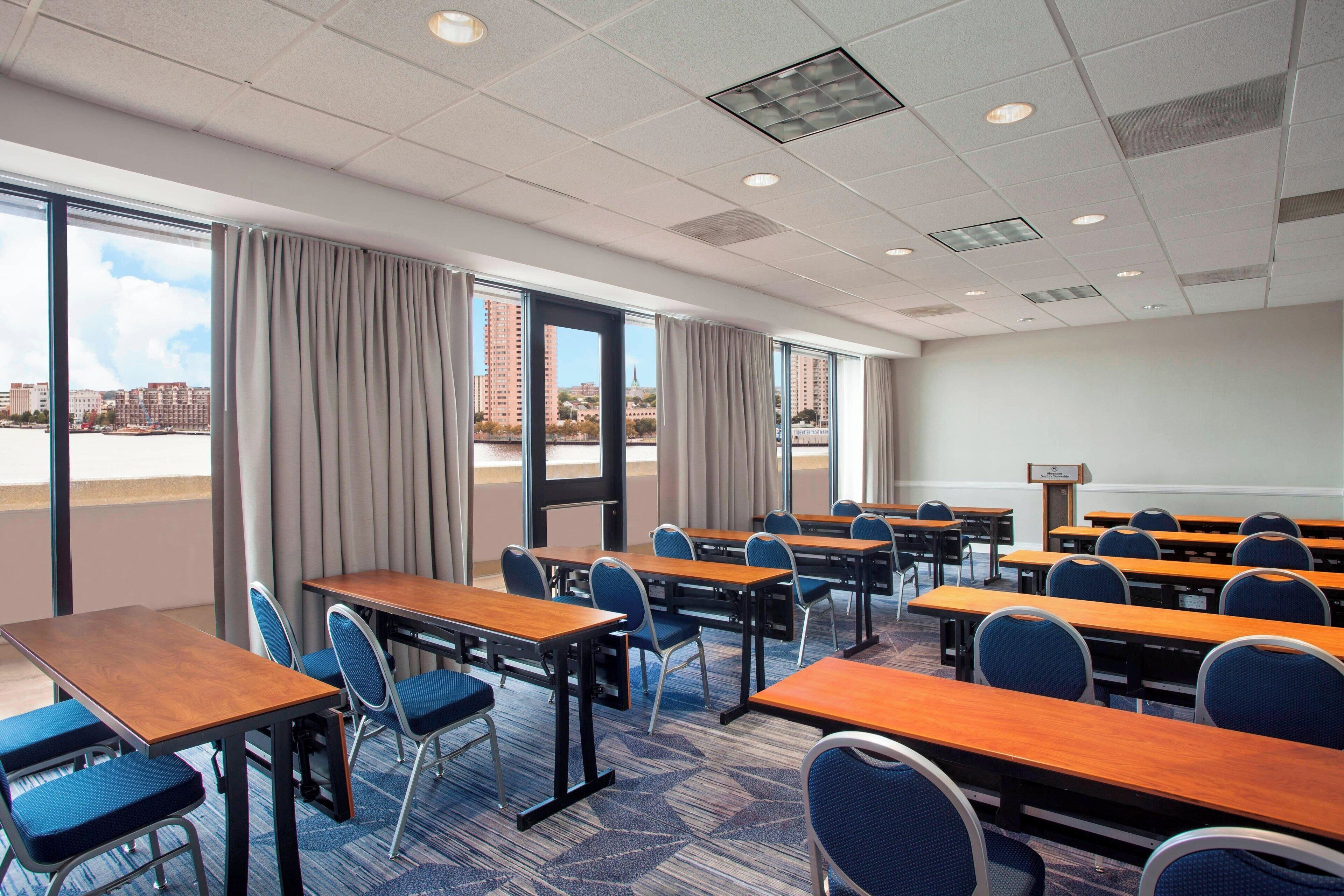 Un salón de conferencias con varias mesas y sillas, ventanas amplias con cortinas y vista a la ciudad.