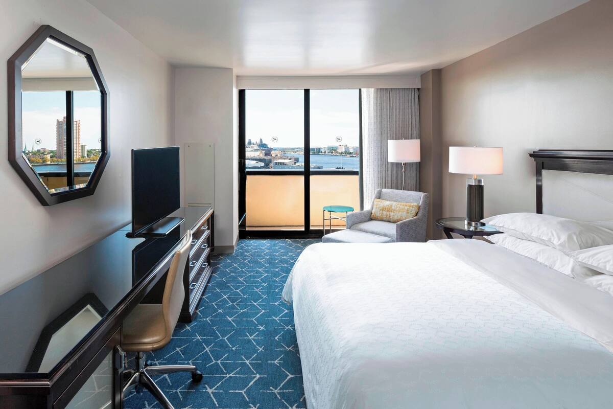 A hotel room with a large bed, a desk with a TV, a chair, a small table with lamps, and a view of a cityscape through large windows.