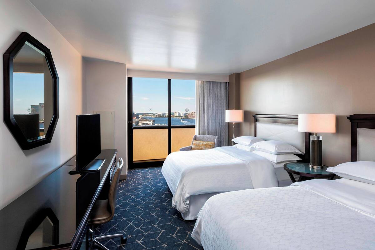 A hotel room with two beds, a desk, a chair, a TV, and a view of a cityscape.