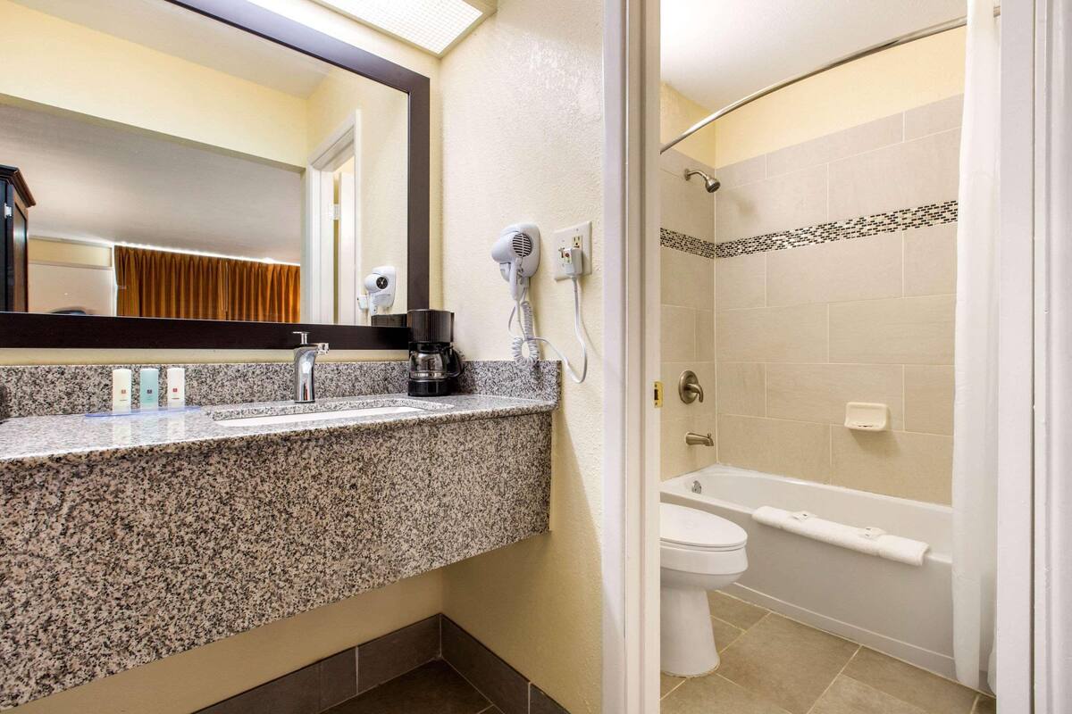A bathroom with a granite countertop, a hairdryer, a coffee maker, a toilet, and a bathtub.