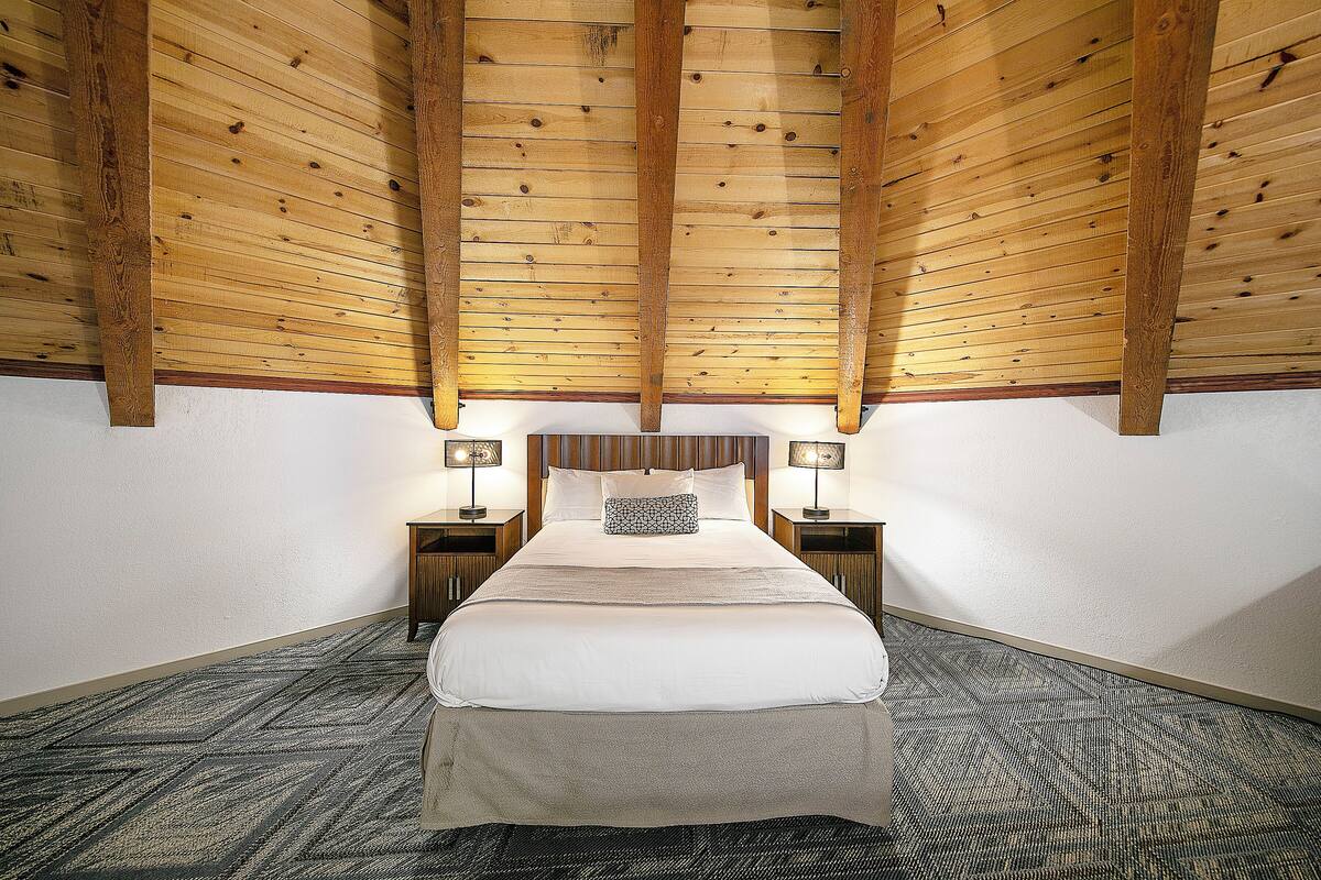 Quarto com teto de madeira, uma cama grande, duas mesas de cabeceira com luminárias e um tapete estampado.