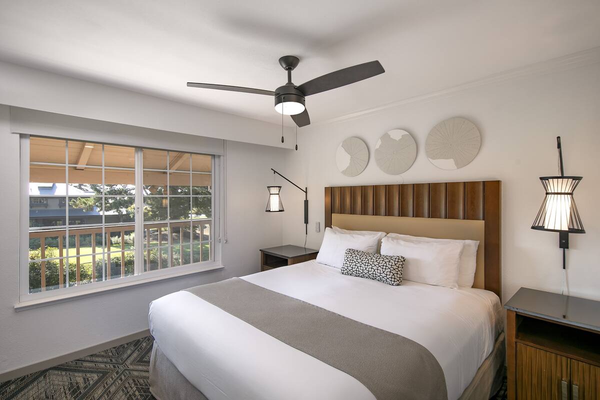 Quarto de hotel com uma cama grande, um ventilador de teto, uma janela com vista para vegetação e uma mesa de cabeceira com abajur.