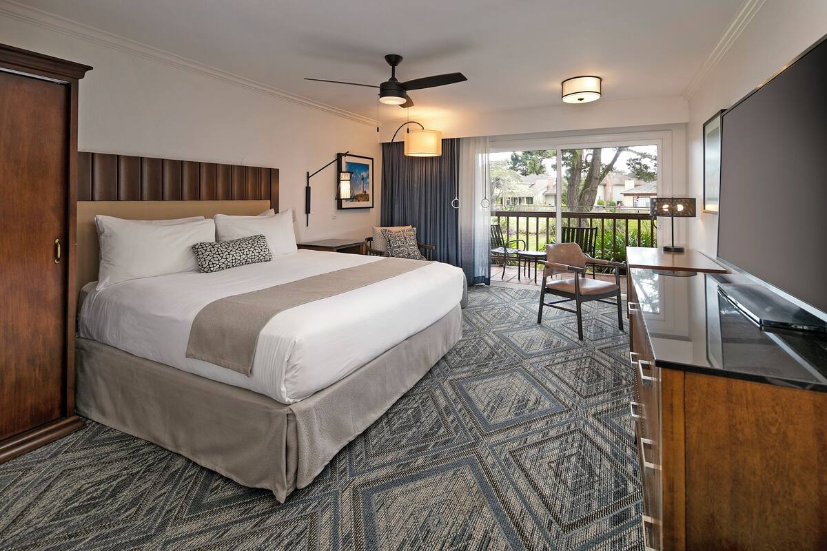 Quarto de hotel com uma cama grande, uma escrivaninha, uma cadeira, um ventilador de teto e vista para o exterior.