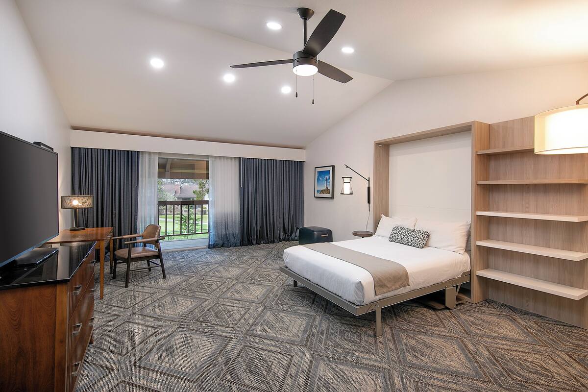 Quarto de hotel moderno com cama, escrivaninha, cadeira e ventilador de teto.