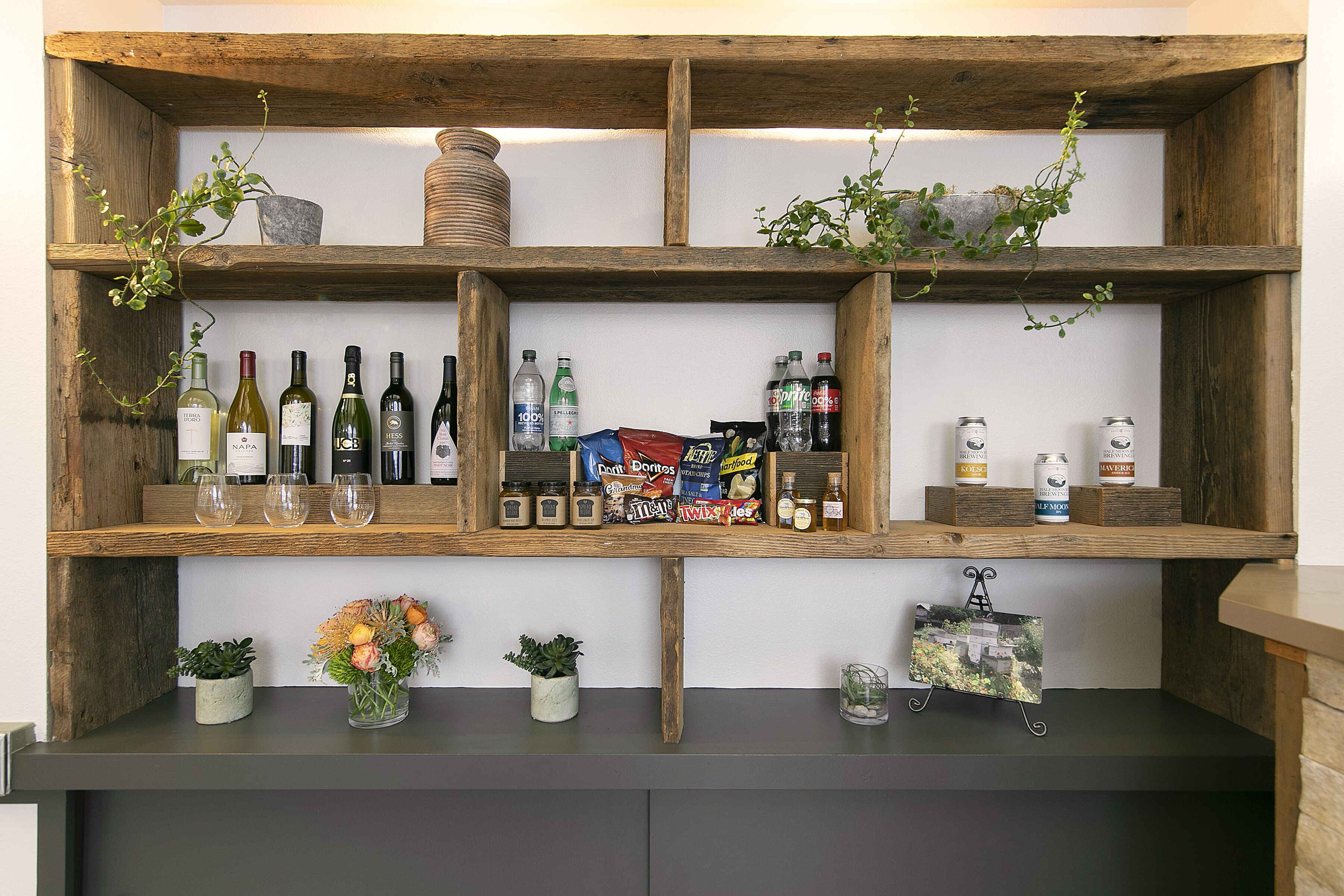 Una estantería de madera con botellas de vino, aperitivos y pequeñas macetas.