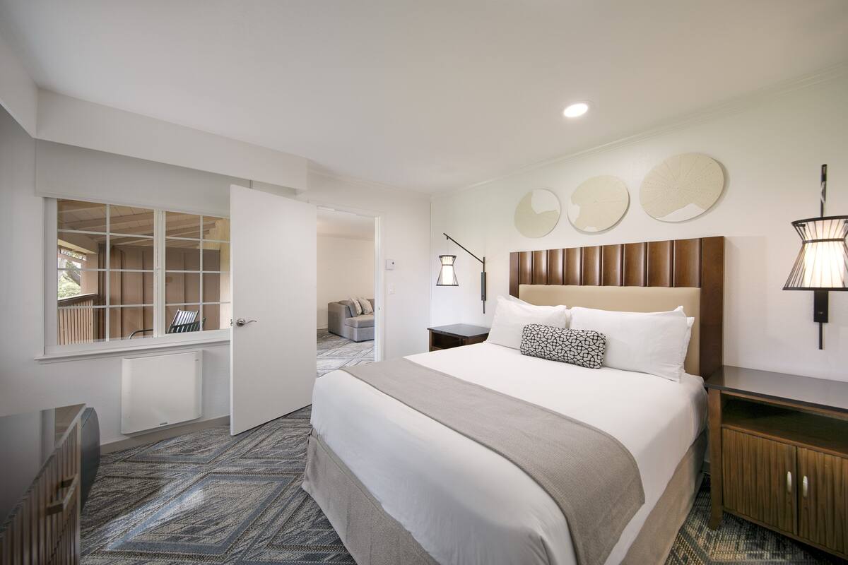 Quarto de hotel com uma cama grande, mesa de cabeceira, luminária e vista para o corredor.