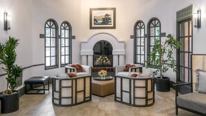 Habitación amplia con techos altos, ventanas grandes y chimenea. Cuenta con varias zonas de estar con cojines y plantas en macetas.