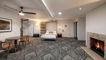 A hotel room with a bed, a desk, a chair, a fireplace, a ceiling fan, and a staircase.