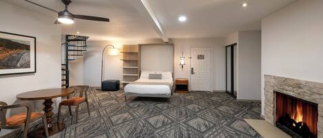 A hotel room with a bed, a desk, a chair, a fireplace, a ceiling fan, and a staircase.
