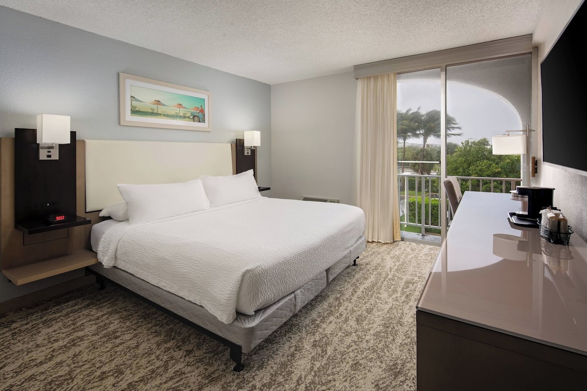 Habitación de hotel con una cama grande, un escritorio y un balcón con vistas a palmeras.