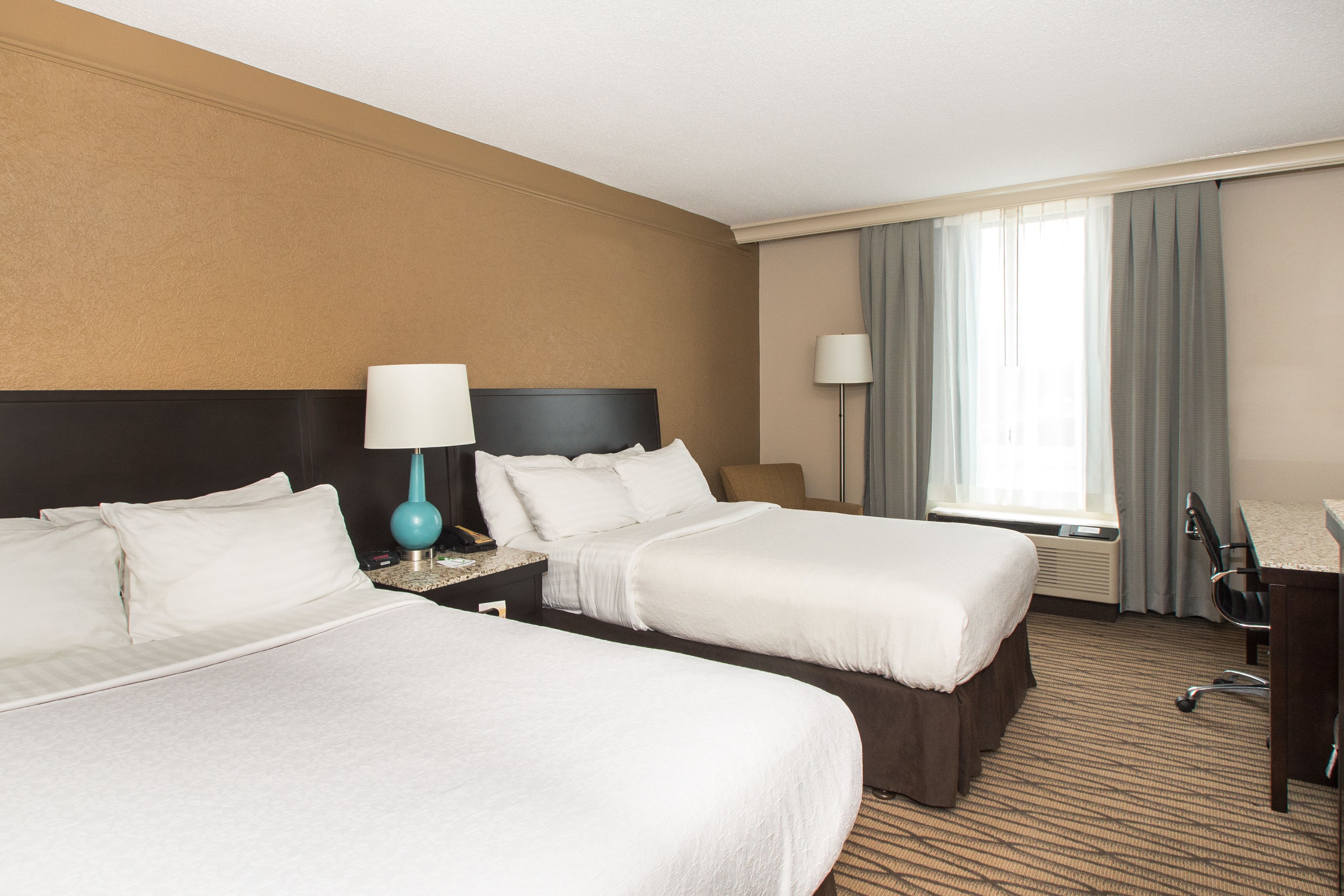 A hotel room with two beds, a desk, a chair, and a lamp.