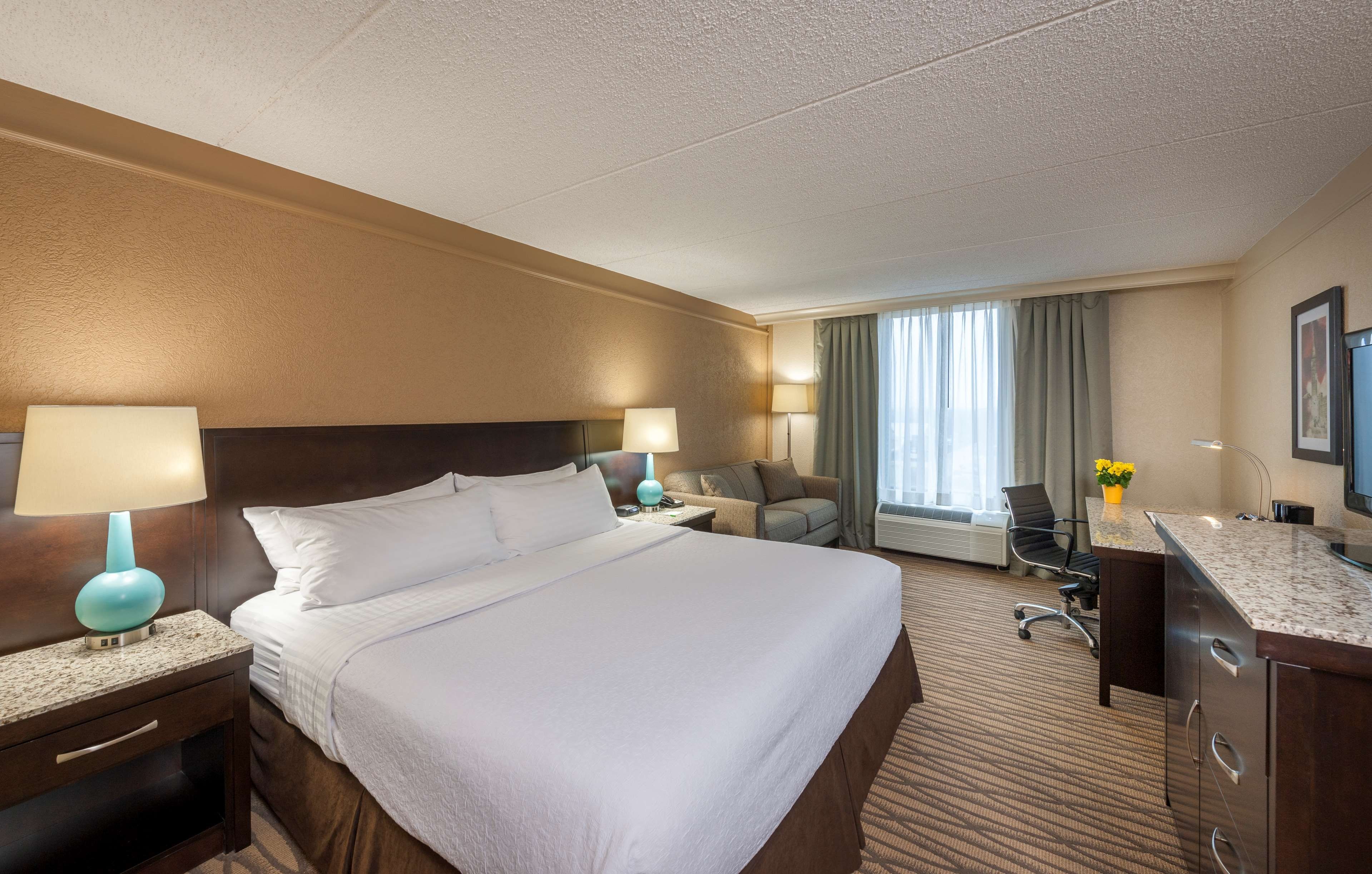 Una habitación de hotel con una cama grande, dos mesitas de noche con lámparas, un escritorio con una silla, un televisor y una ventana con cortinas.