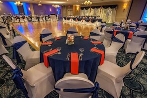 A banquet hall with round tables set for dining, white chairs with blue sashes, and a stage with draped curtains in the background.