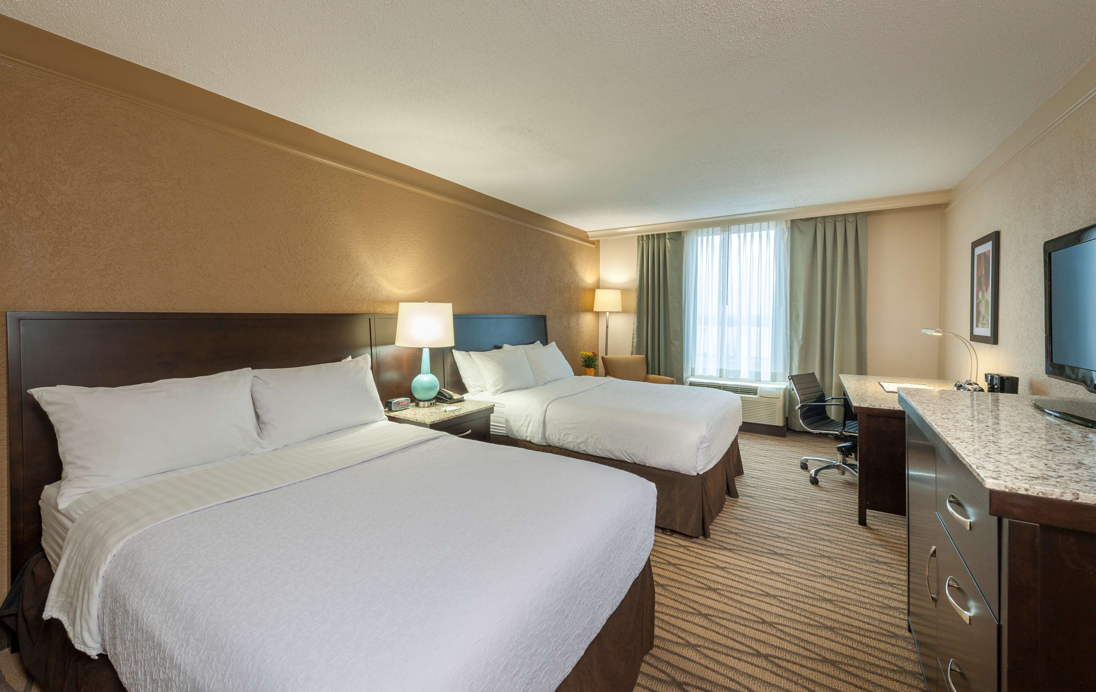 Une chambre d’hôtel avec deux lits, un bureau, une chaise, une télévision et une fenêtre avec des rideaux.