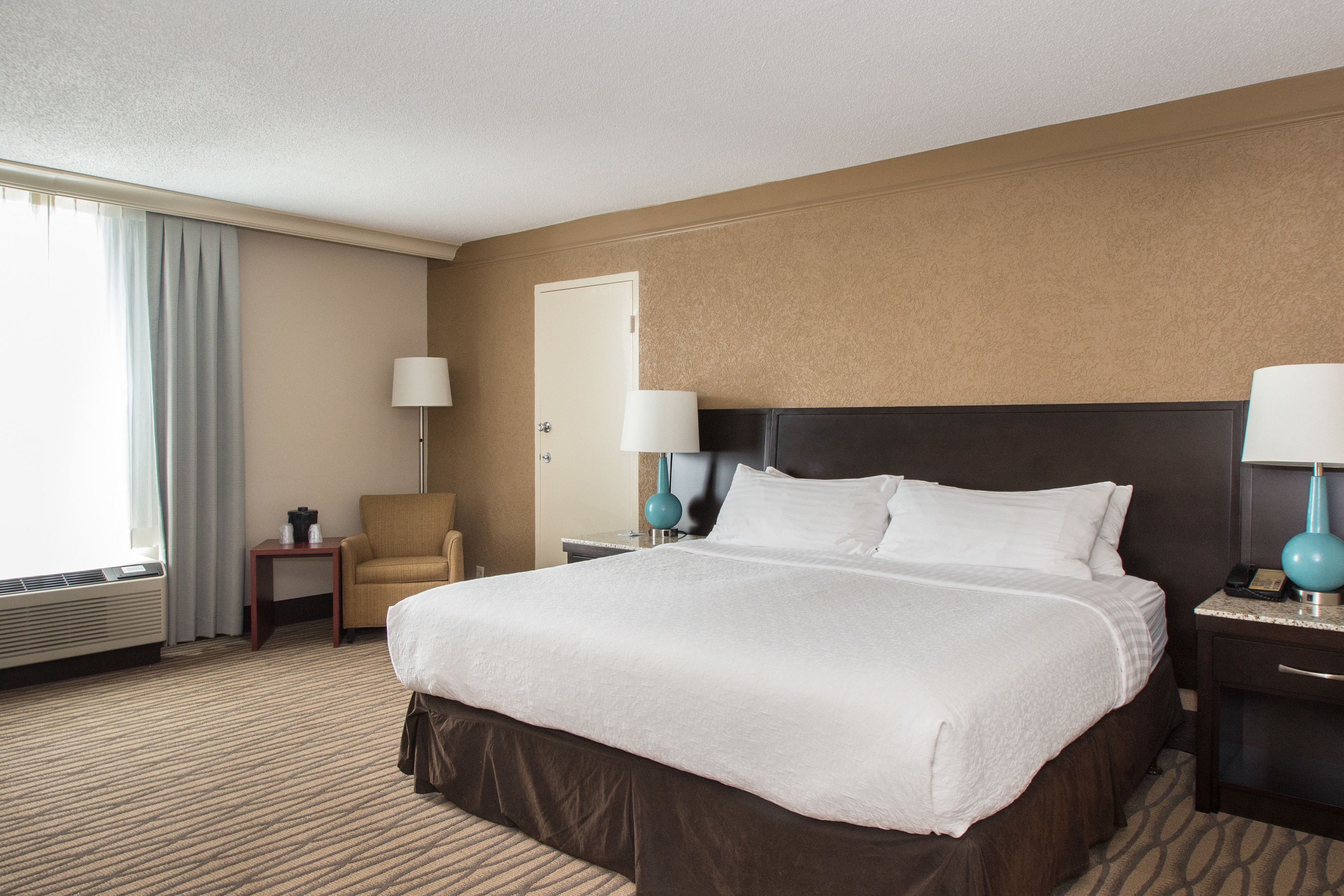 A hotel room with a large bed, two bedside tables with lamps, a chair, a desk, and a door.