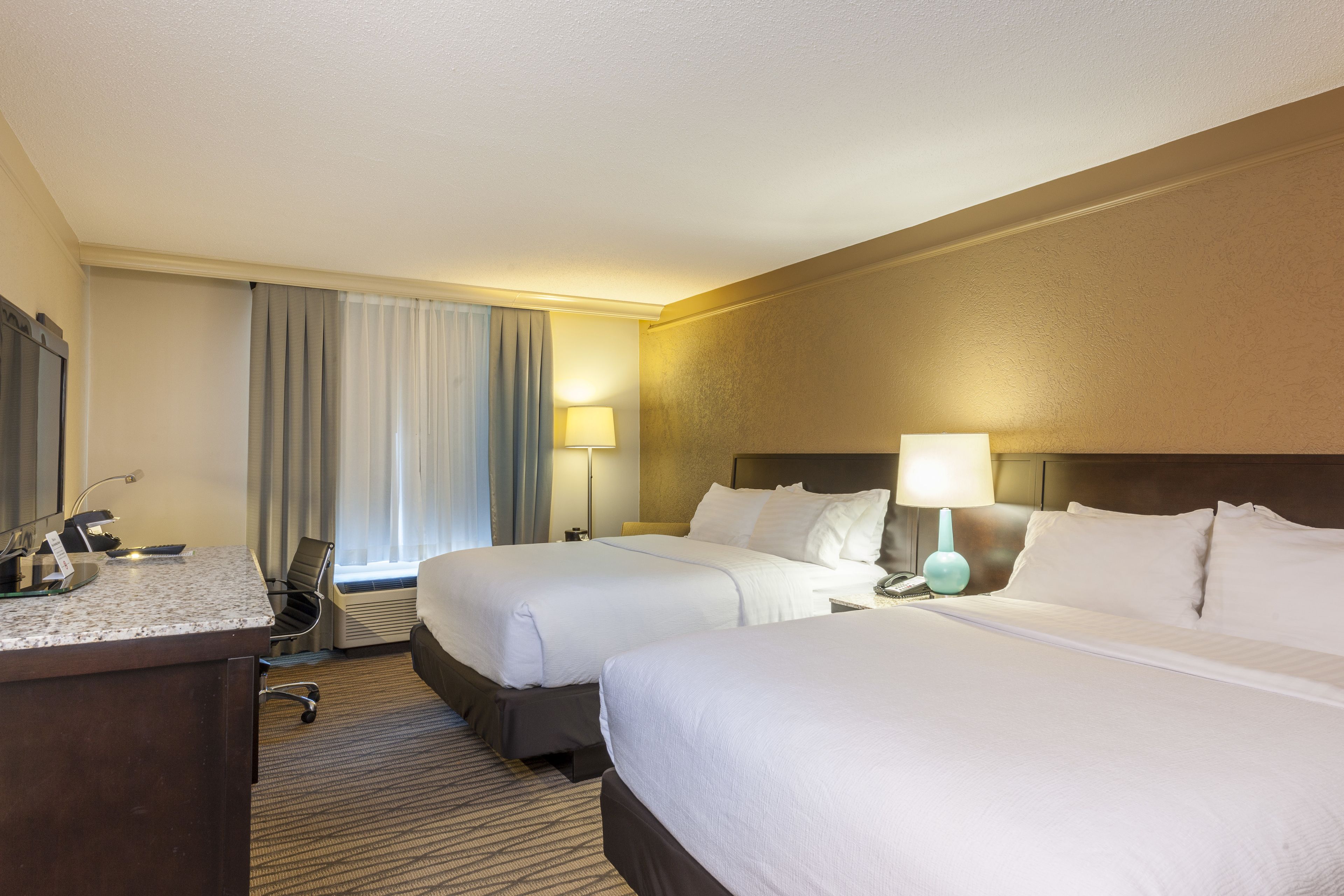 A hotel room with two beds, a desk, a chair, a lamp, and a window with curtains.