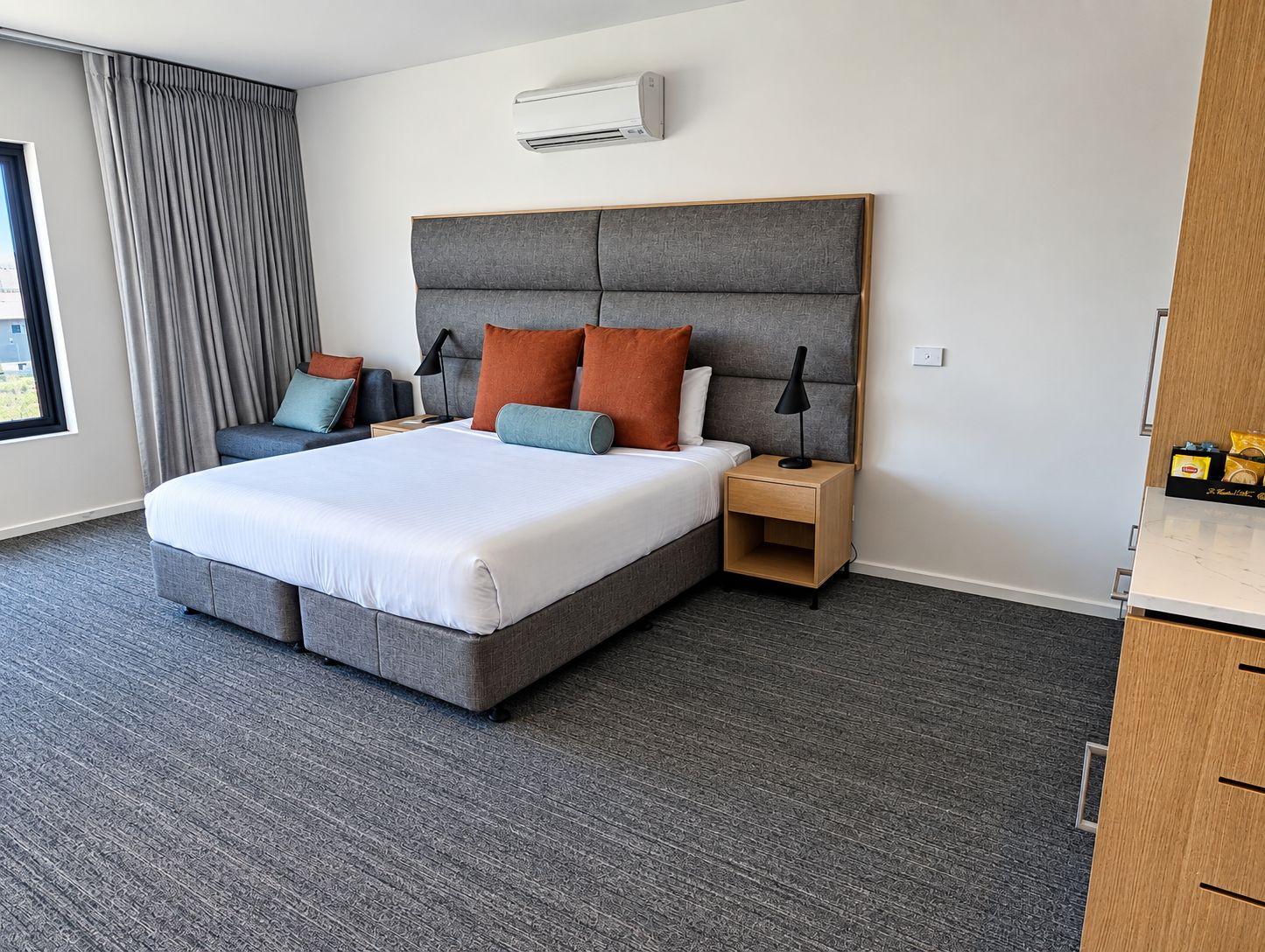 Una habitación de hotel moderna con una cama grande, cabecero tapizado en gris y una mesita de noche con lámpara.