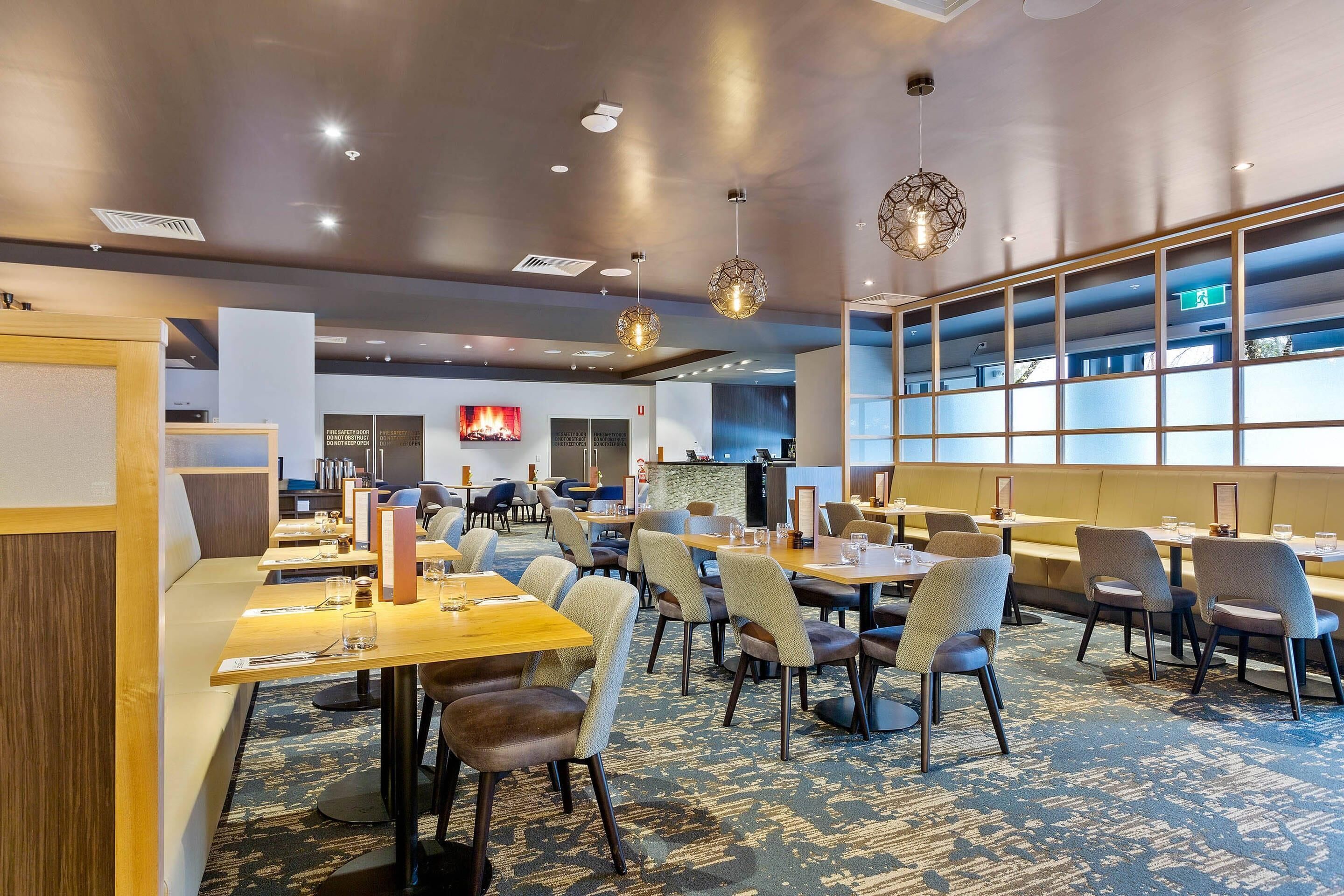 A modern hotel dining area with multiple tables and chairs, a fire feature, and a view of the city through large windows.
