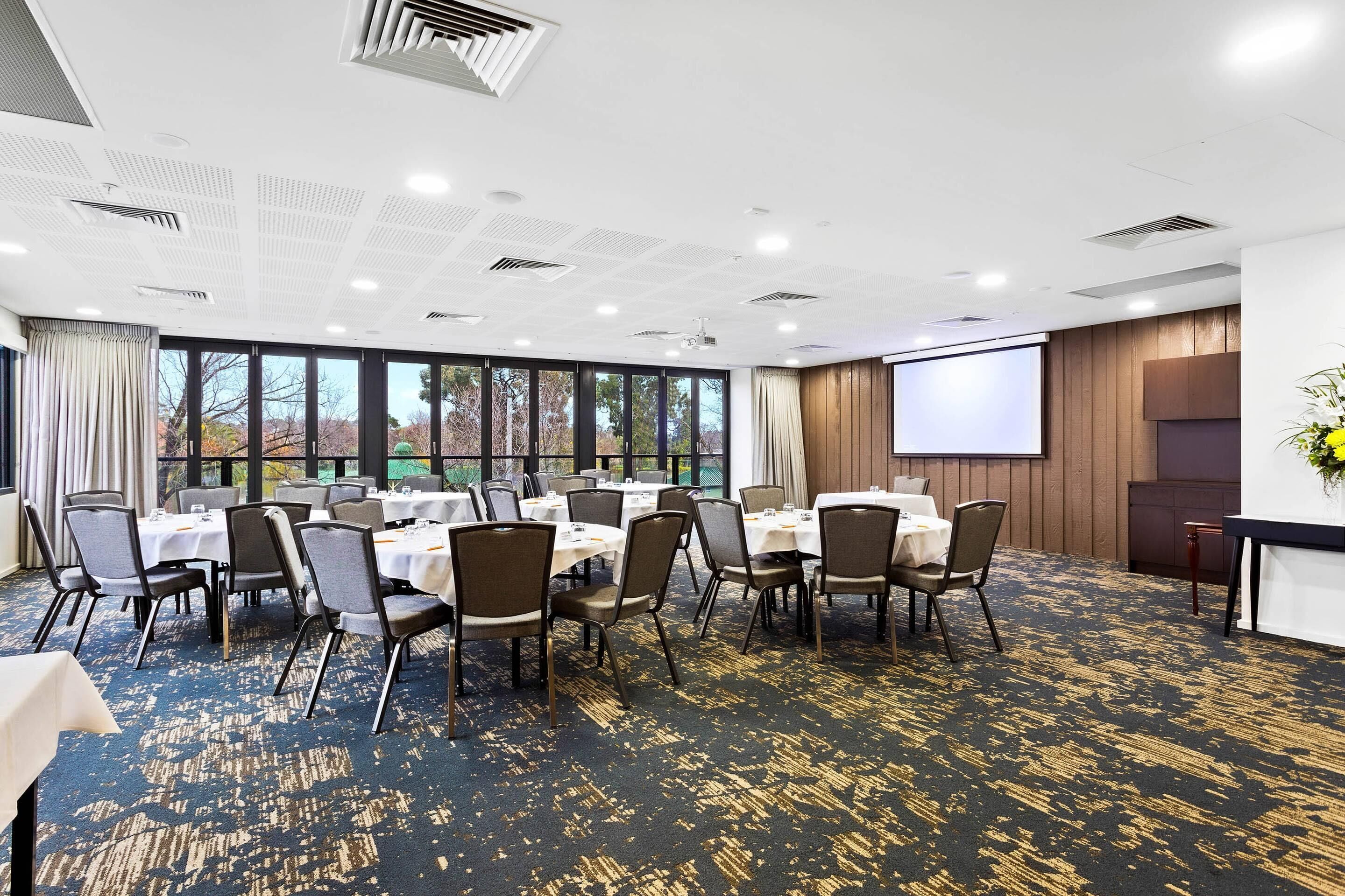 Un salón de conferencias con mesas y sillas, amplias ventanas, una pantalla de proyector y piso alfombrado.