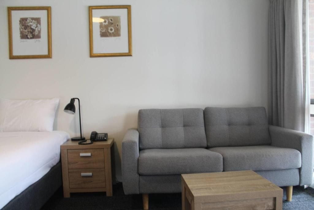 Habitación de hotel con cama, sofá gris, mesita de noche de madera con lámpara y dos cuadros en la pared.