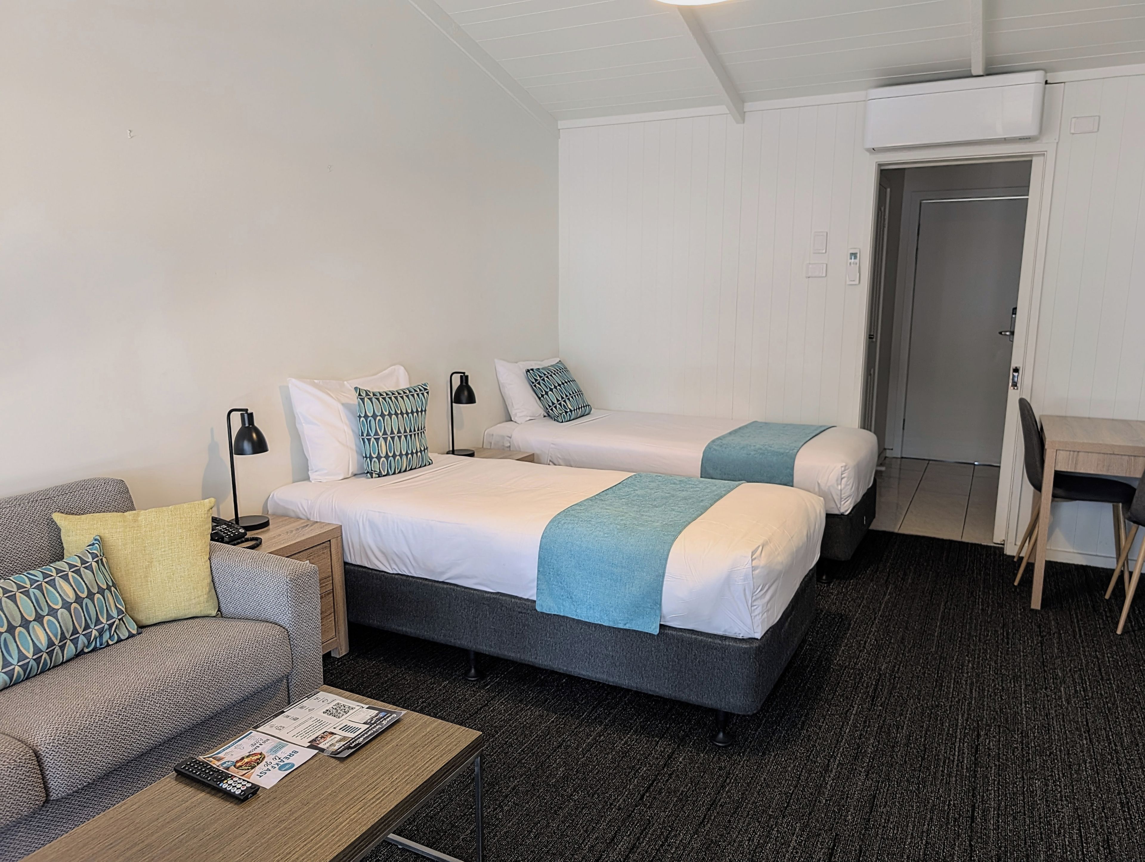 Ett hotellrum med två separata sängar, en soffa, ett litet bord med ett tidningsnummer och ett skrivbord med en stol.