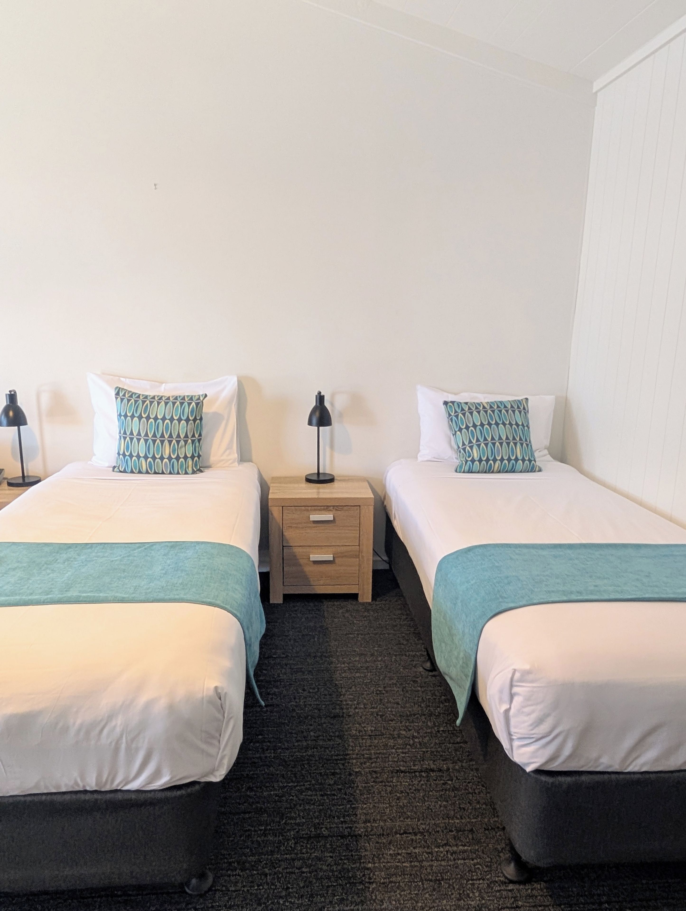 A hotel room with two beds, each with a patterned pillow, a bedside table with a lamp, and a dark carpet.