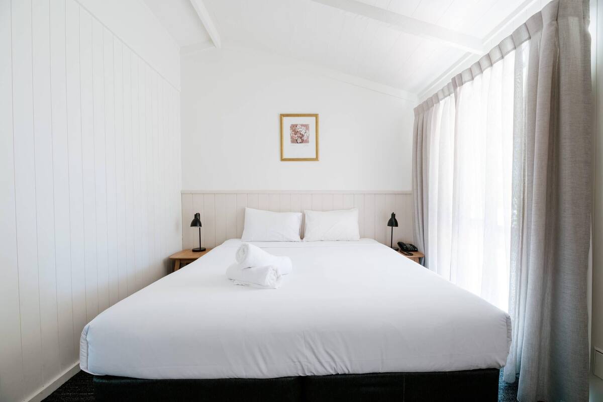 Ein Hotelzimmer mit einem großen Bett, weißen Wänden, einem gerahmten Bild an der Wand und einem Fenster mit Vorhängen.