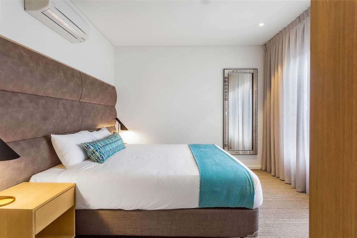 Ein Hotelzimmer mit einem großen Bett, einem Nachttisch, einem Spiegel und Blick durch Vorhänge nach draußen.