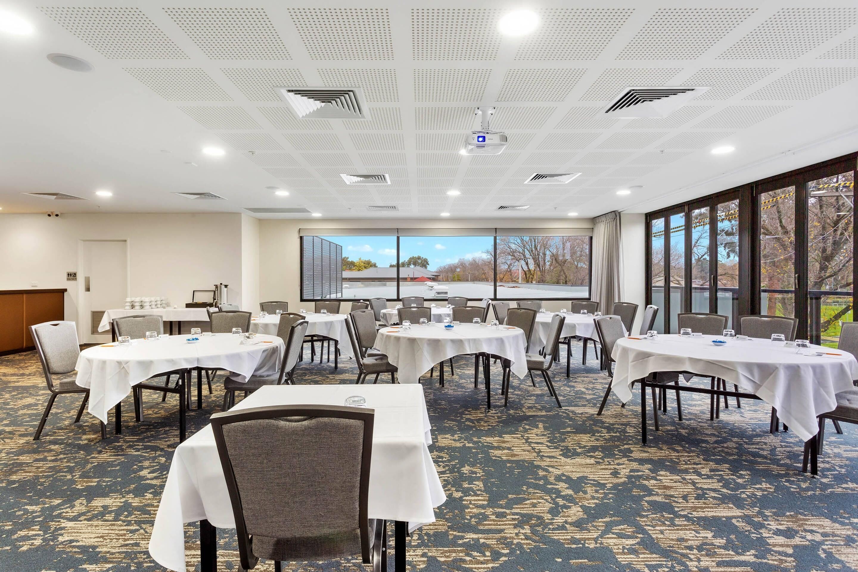 Un salón de conferencias con varias mesas y sillas, amplias ventanas con vistas a árboles, e iluminación empotrada y un proyector en el techo.