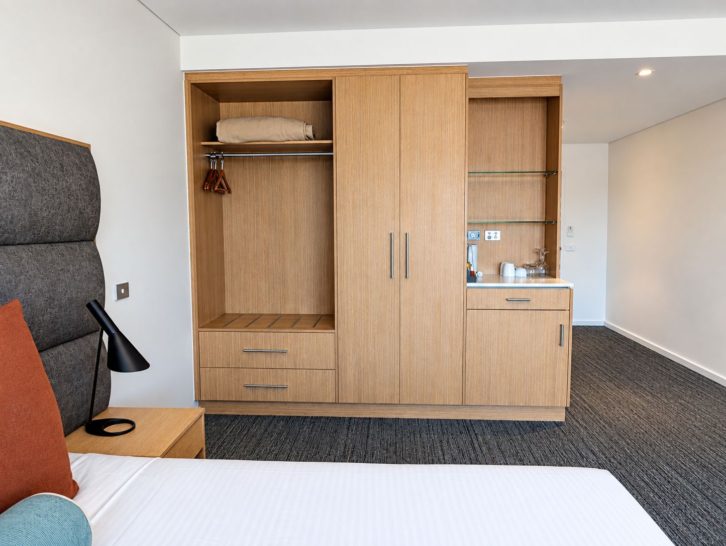 A hotel room with a bed, a wardrobe with a mirror, a small kitchenette, and a bedside table with a lamp.