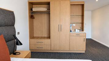 A hotel room with a bed, a wardrobe with a mirror, a small kitchenette, and a bedside table with a lamp.