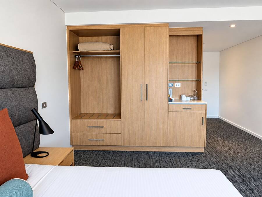 Une chambre d’hôtel comprenant un lit, une armoire avec un miroir, une petite kitchenette et une table de chevet avec une lampe.