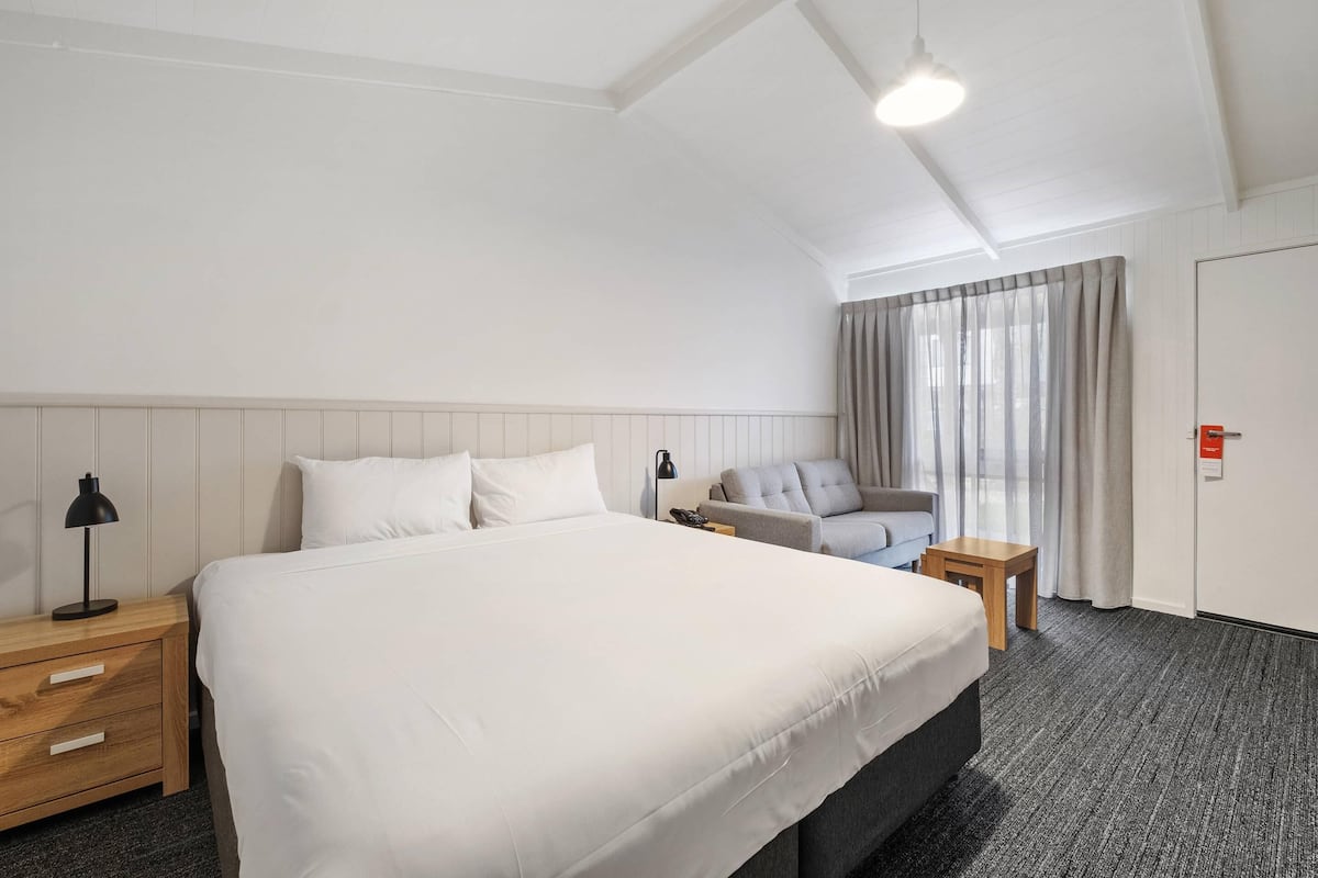 Ein Hotelzimmer mit einem großen Bett, einem Nachttisch mit Lampe, einem kleinen Tisch, einem Sessel und einem Fenster mit Vorhängen.