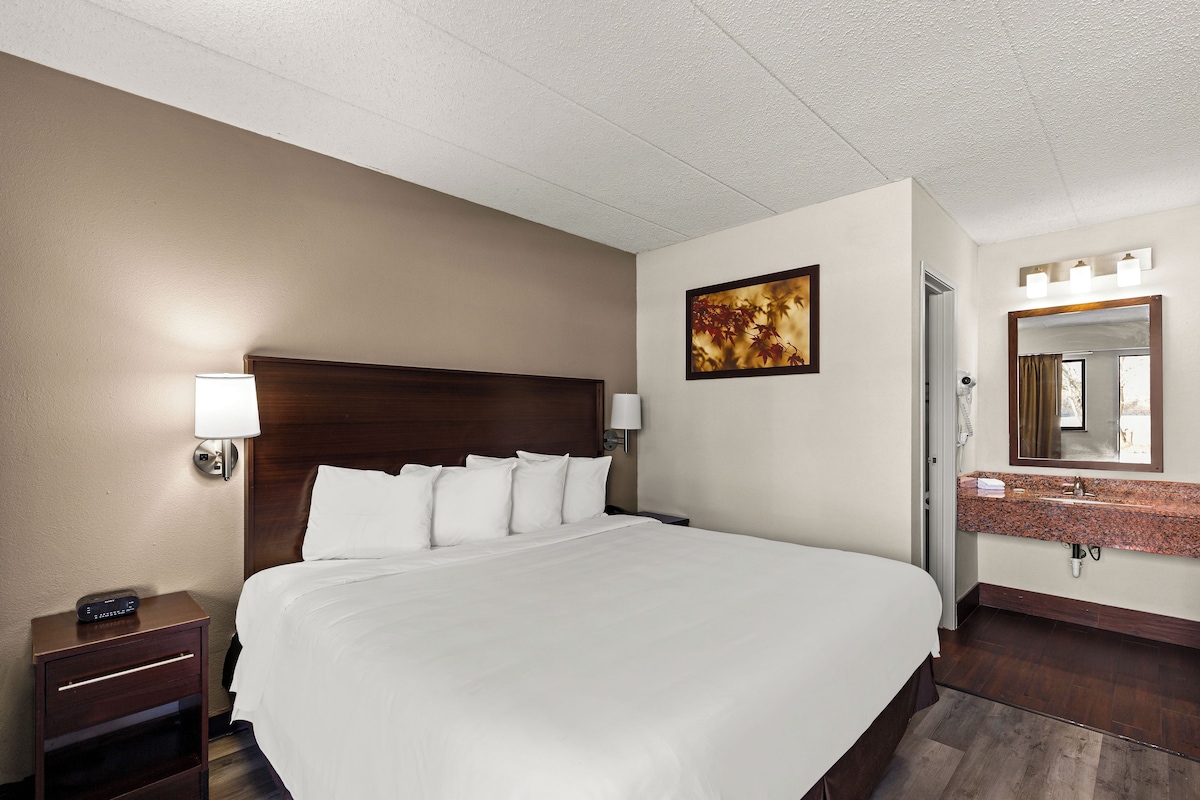 A hotel room with a large bed, a nightstand with a lamp, a painting on the wall, and a bathroom with a sink and mirror.