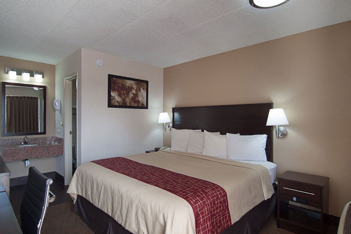 A hotel room with a bed, bedside lamps, a nightstand, a desk with a chair, a bathroom mirror, and a framed picture on the wall.